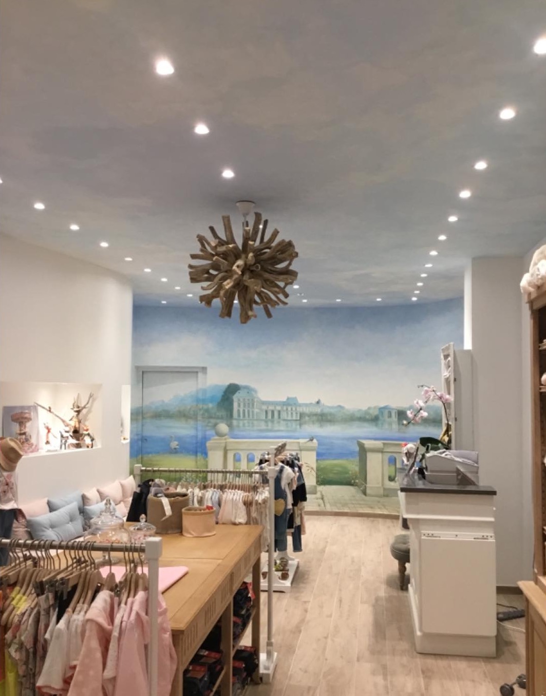 L'intérieur de la boutique : fresque murale de château sous un ciel peint, lustre en bois flotté et portants d'enfants